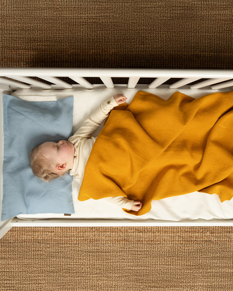 Baby sleeping in a white crib under a mustard yellow linen blanket, on a natural brown woven rug.