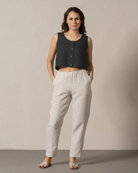 A woman with dark hair wearing the menique 100% linen pants and linen crop top set. Her hands are in the pockets, while she is looking stright. The background of the photo is bright and in cool tones.