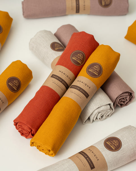 A top-down product shot featuring several rolls of textured linen fabric scattered across a clean white surface. The fabrics come in rich, earthy colors including mustard yellow, rust orange, light beige, and dusty mauve. Each roll is secured with a brown kraft paper sleeve printed with the brand name "MENIQUE" and "PREMIUM QUALITY," along with a round brown sticker that reads "MADE FROM 100% LINEN." A rust orange and a mustard yellow roll are prominently crossed in the center of the composition.