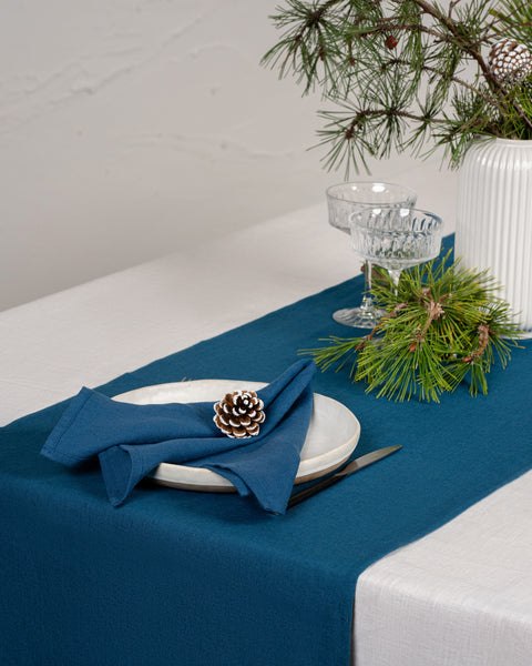 Deep teal linen table runner with matching linen napkin on a white tablecloth, styled with pine branches and winter holiday décor.