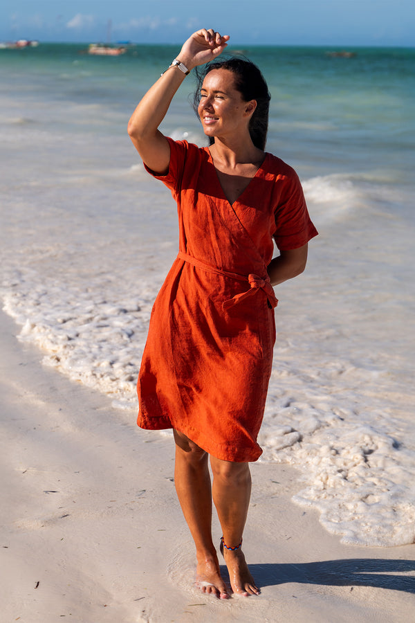 A woman with dark hair smiles and shields her eyes with one hand as she stands barefoot on a sandy beach. She is wearing a cinnamon red linen wrap dress with short sleeves. The ocean is in the background with small waves and a few boats visible in the distance under a bright sky.