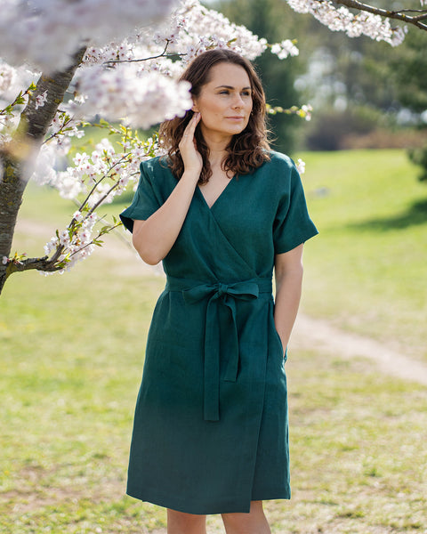 A woman with brown hair, wearing a dark green, short-sleeved wrap dress, stands outdoors under a tree with white blossoms.
