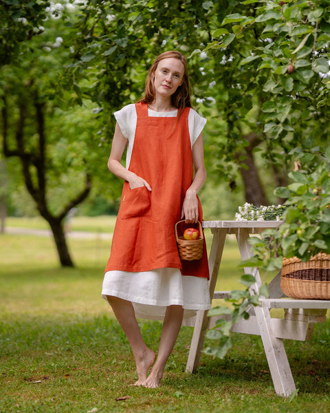 A woman with long reddish-brown hair, wearing a menique linen apron, stands barefoot in a tranquil garden, holding a basket of red apples.