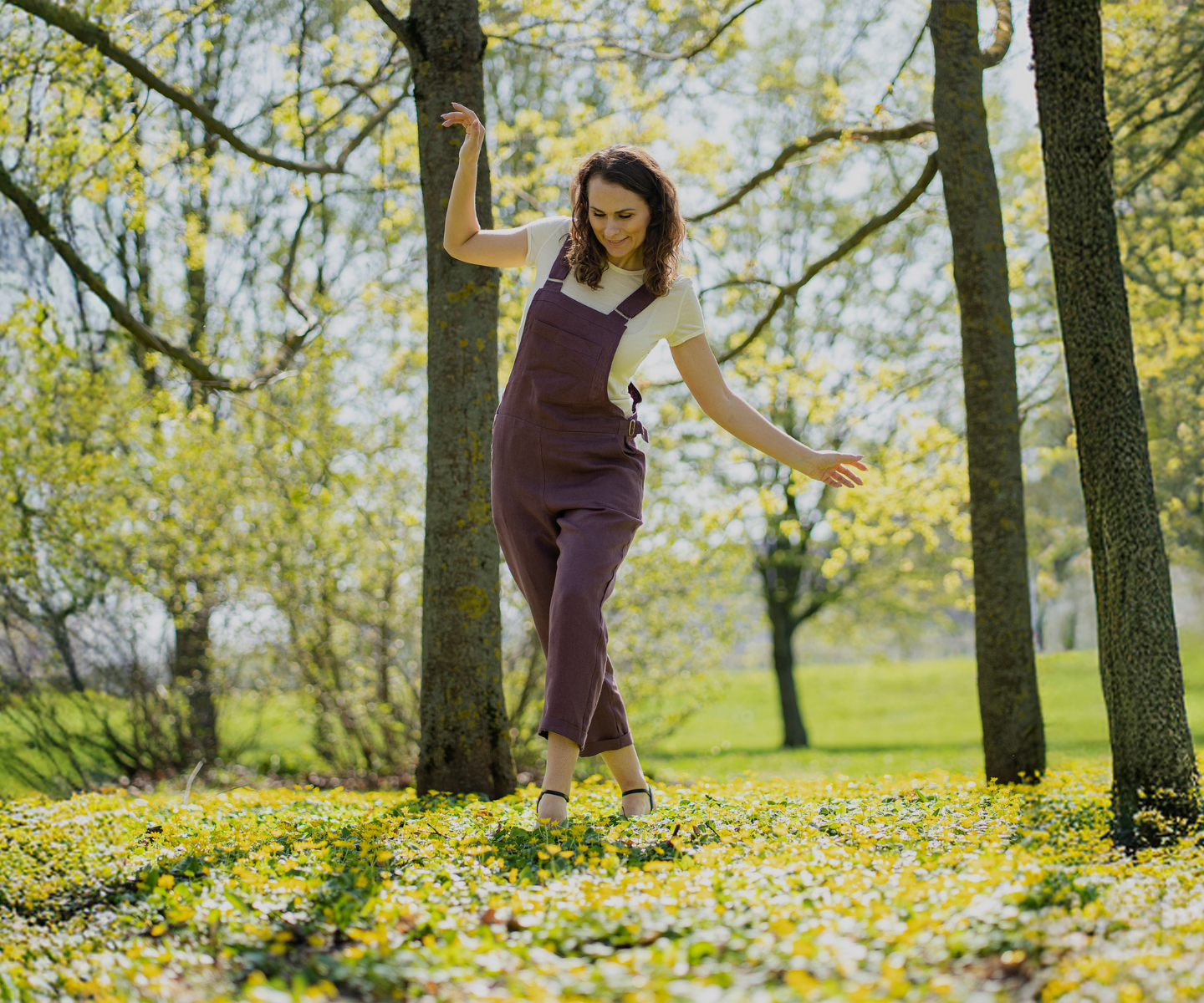 Woman wearing purple sleeveless jumpsuit layered over white t-shirt, relaxed fit women’s overalls style, breathable natural fabric one-piece outfit styled outdoors in spring nature setting