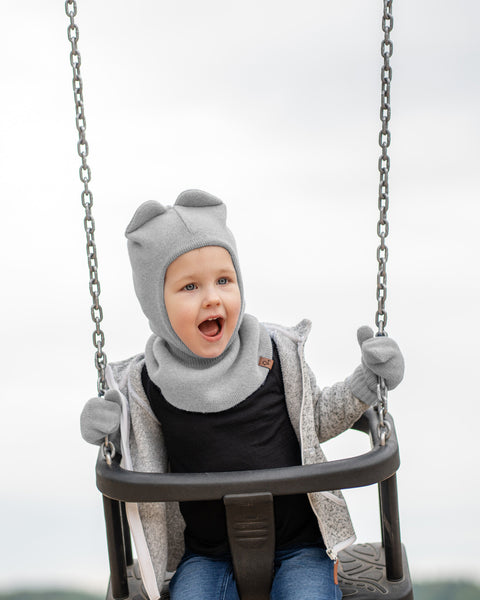 A young child, appearing to be a toddler, is smiling widely while sitting in a swing. They are wearing a light grey hat with what look like small ears on top, a grey knitted neck warmer, and grey mittens. Underneath, they have on a dark shirt and blue jeans. The chains of the swing are visible on either side, and the background is a light, overcast sky.