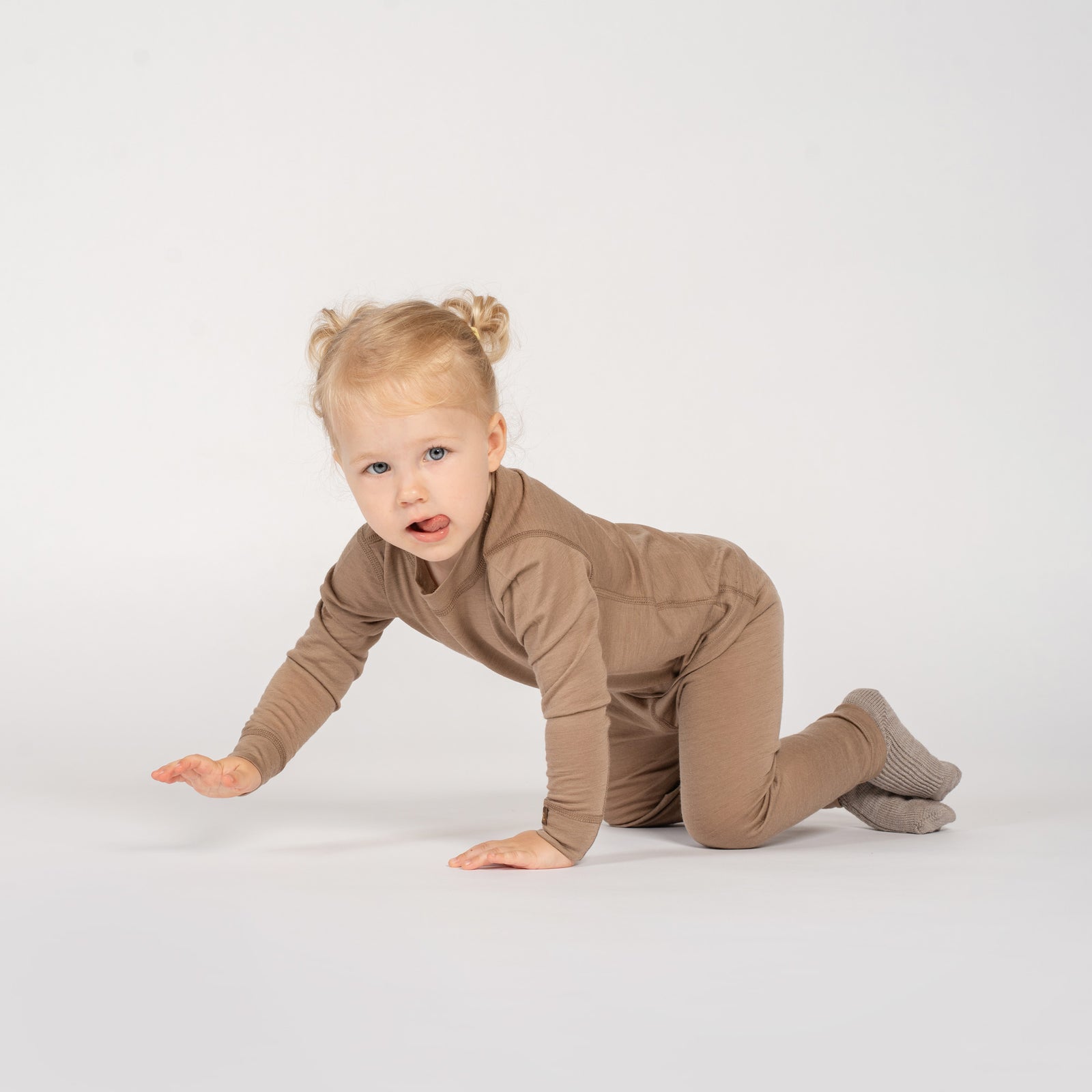 A smiling toddler stands outdoors in a lush, green forest setting, looking down gently at the ground. The child is wearing a light brown long-sleeve top paired with olive green shorts that feature a small acorn patch on the pocket.
