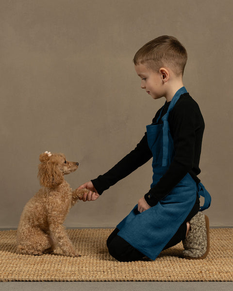 Child in a cobalt blue apron kneeling on a woven mat with a small brown dog.