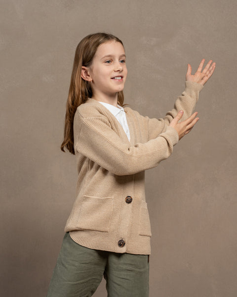 A girl wearing the menique merino wool cardigan for kids in beige color. She is swinging her hands from side to side, emphasizing the length of the sleeves. The cardigan also has front pockets on both sides, and a button closure.