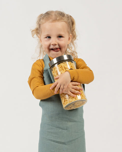 A young child with light curly hair, pulled into ponytails, is standing and holding a large glass jar filled with yellow, flaky cereal. The child is looking at the viewer with a slight smile and their tongue sticking out a little. They are wearing a long-sleeved spicy yellow shirt and a mint green apron over it. The background is white.