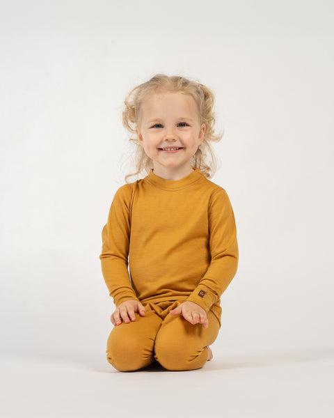 A happy child with curly blonde pigtails, wearing a yellow long-sleeved top and matching pants, is kneeling on a white surface and smiling at the camera. Their hands are resting on their lap.