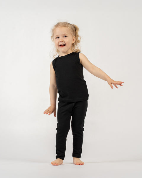 A young child with light curly hair in pigtails is standing barefoot against a plain white background. They are wearing a black sleeveless top and black leggings. The child is looking towards the viewer with an open-mouthed smile, and their arms are outstretched slightly to the sides, as if in mid-movement or about to dance.