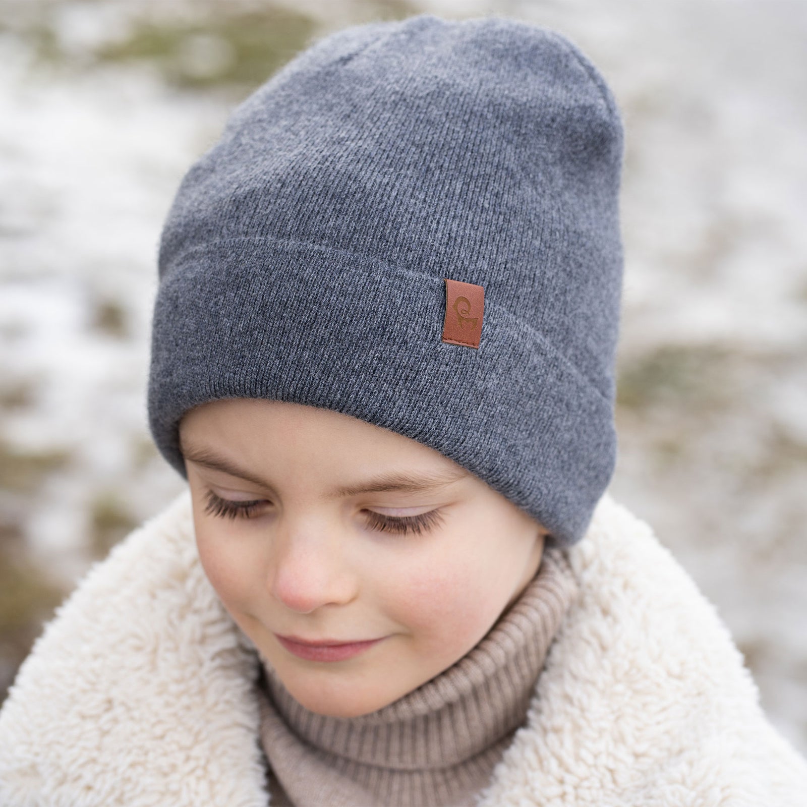 A young child with short brown hair and a wide smile, is dressed in winter clothing. They are wearing a beige beanie, a beige turtleneck sweater, and a cream-colored sherpa jacket, with beige knitted gloves. A snowy, blurred background with what appears to be a body of water is visible behind them.