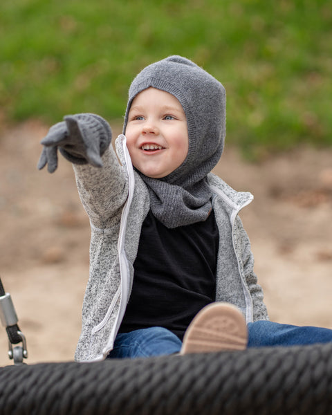 A young child with blue eyes and a wide smile, is sitting on a black rope swing, pointing to the left with their arm outstretched. They are wearing a dark grey balaclava, a grey speckled zip-up hoodie, dark grey mittens, blue jeans, and light pink sneakers. The background is a blurred outdoor scene with sand and grass.
