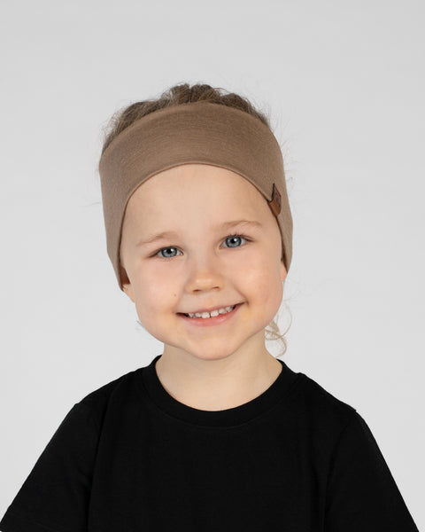 A close-up studio portrait of a smiling young child looking directly at the camera against a plain light grey background. She is wearing a black short-sleeved t-shirt and a wide beige headband with a small brown logo tag on the side.