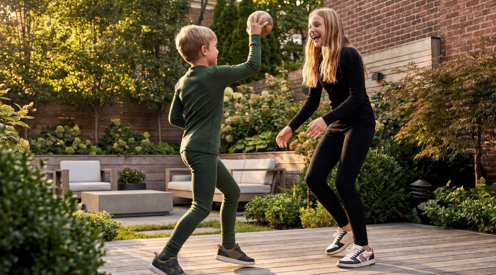 A boy and an older girl happily playing catch with a ball on an outdoor wooden deck. The boy is wearing a dark green long-sleeve top with matching green pants, and the girl is wearing a black long-sleeve top with black leggings.