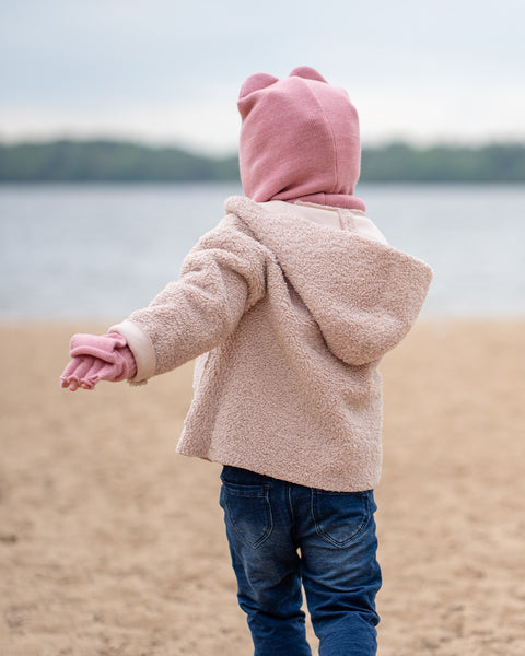 A young child, seen from behind, wears a pink balaclava with ears, a beige coat, and blue jeans while standing on a sandy beach looking out at the water.