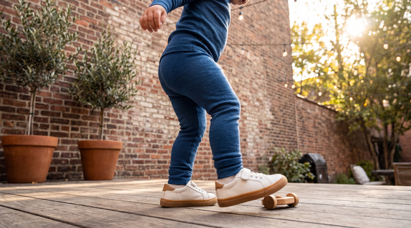 A close-up shot focusing on the legs and lower torso of a young child stepping on a wooden deck. The child is wearing blue fitted pants and white sneakers with velcro straps, with a small wooden toy visible near their feet.