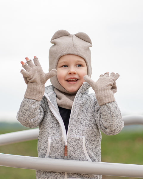 A young child with blonde hair and blue eyes, wears a creamy beige balaclava with ears, a matching scarf, gloves, and a grey speckled zip-up hoodie. 