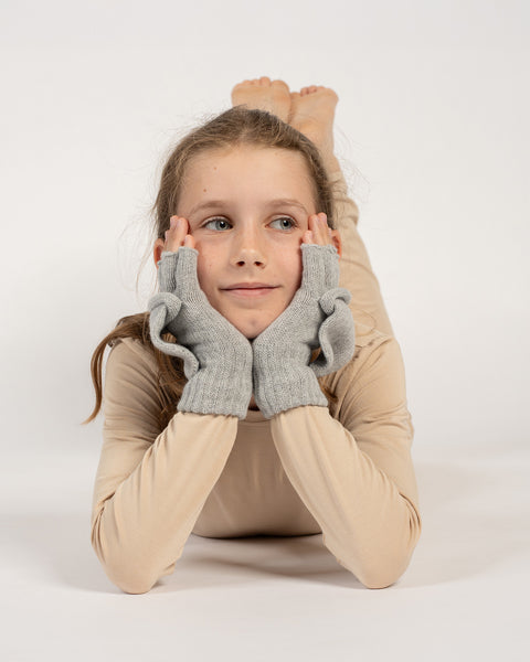 Girl lying down with head resting on hands, wearing light gray knitted convertible mittens with mitten flaps open.