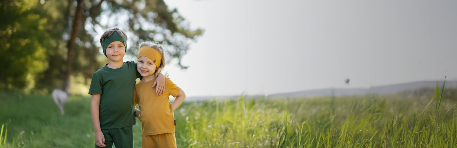 A young boy in a dark green outfit and matching headband standing with his arm warmly around a young girl in a mustard yellow outfit and headband in a bright, grassy field.