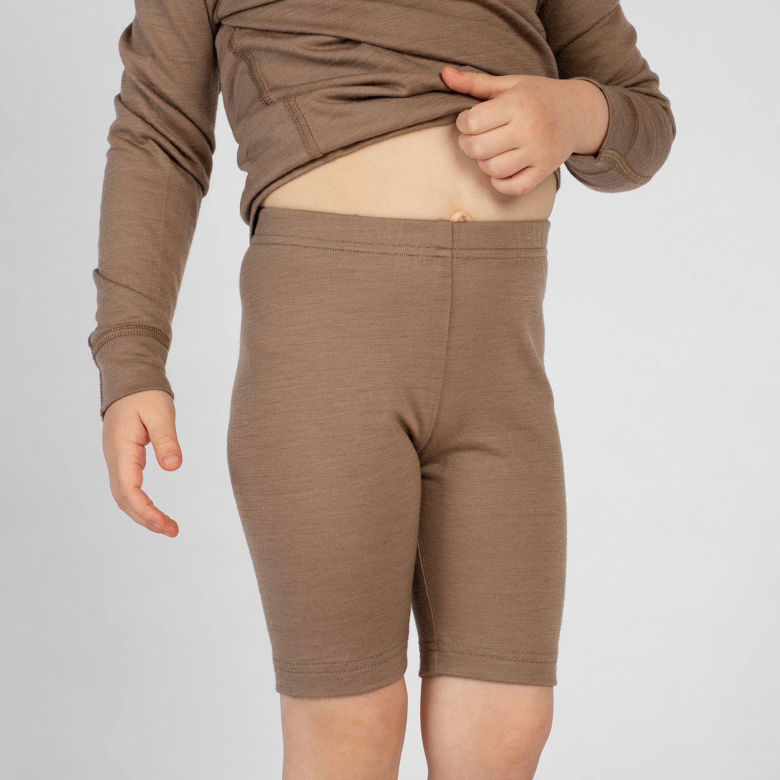 A slightly angled view of a child wearing dark charcoal merino wool cycling shorts paired with a brown long-sleeve top. The child is lifting the front of the shirt with one hand to display the high-waisted fit of the shorts, set against a plain light gray studio background.