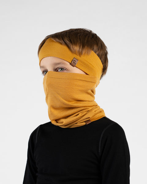 A close-up studio portrait of a young boy looking off to his left in a three-quarter profile against a white background. He is wearing a black long-sleeved top, a mustard yellow headband, and a matching mustard yellow neck gaiter pulled up over his nose and mouth.