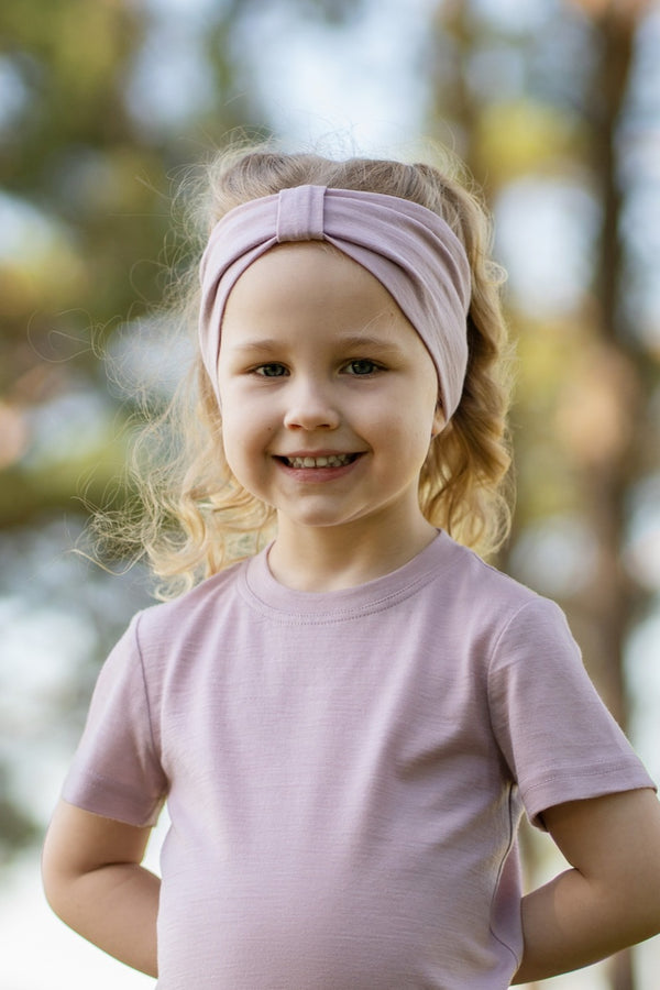 Young girl smiling outdoors wearing a dusty pink Merino wool T-shirt and a matching Merino wool headband. The soft, breathable accessories and clothing are showcased in a sunny natural setting, offering comfortable everyday wear for active ki