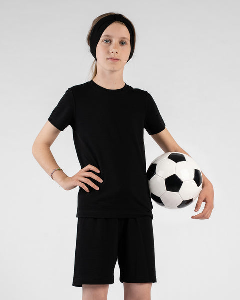 A studio portrait of a young girl standing with one hand on her hip, looking directly at the camera against a white background. She is holding a black and white soccer ball under one arm and wearing a black short-sleeved t-shirt, black shorts, and a black ribbon headband.
