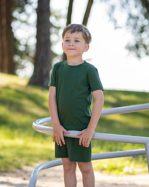 Young boy smiling outdoors wearing a dark green Merino wool T-shirt with a matching Merino wool shorts. The soft, breathable outfit and accessory are showcased in a sunny natural setting, offering comfortable everyday wear for active kids.