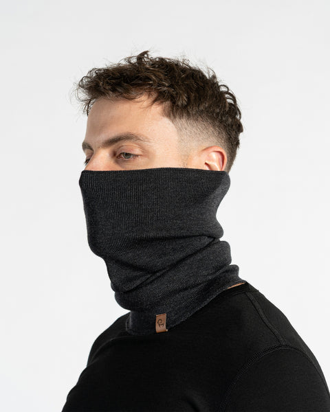 A man with short, dark, wavy hair, wearing a dark gray knit neck gaiter pulled up to cover his nose and mouth, and a black long-sleeved shirt. He is looking to his right. A small brown leather tag is visible on the lower front of the gaiter. The background is plain white.