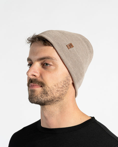 A man with light skin, blue eyes, and a beard. He wears a creamy beige colored menique knit beanie with a small brown patch on the side, and a black crew-neck shirt.