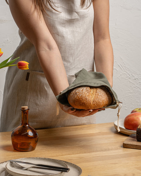 A person wearing a natural color linen apron holds a round loaf of bread inside a stone green menique linen drawstring bag over a wooden table. Tulips and apples are nearby.