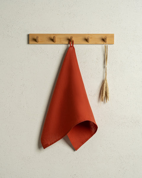 The menique linen kitchen dish towel in the color of cinnamon red hanging on a wooden rack on a wall.