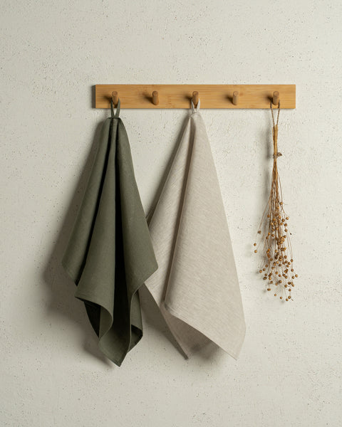 Two menique linen kitchen dish towels hanging on a wooden rack on a wall. The colors of the towels are stone green and natural. 