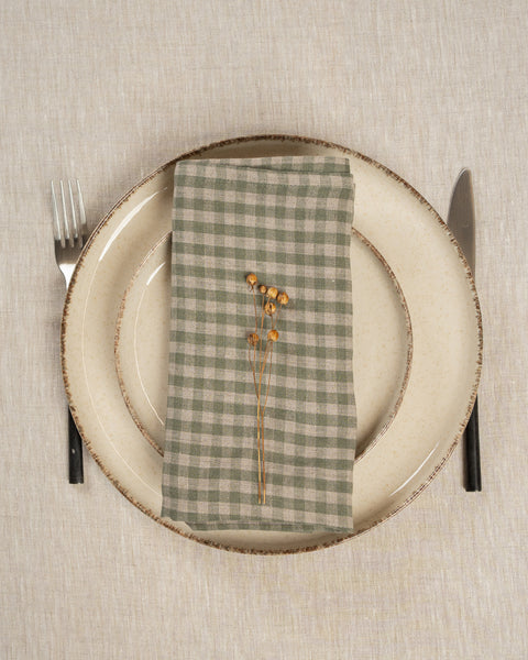 The menique linen napkin in green check color, put on top of two stacked plates . There are cutlery items by the sides of the plates. The background is a natural color linen tablecloth.