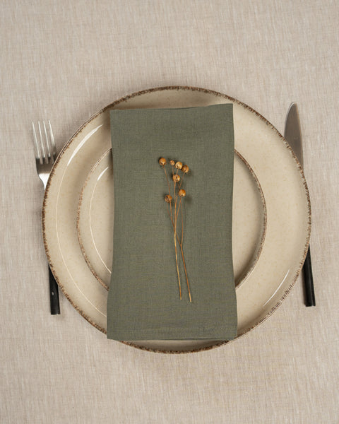 The menique linen napkin in stone green color, put on top of two stacked plates . There are cutlery items by the sides of the plates. The background is a natural color linen tablecloth.