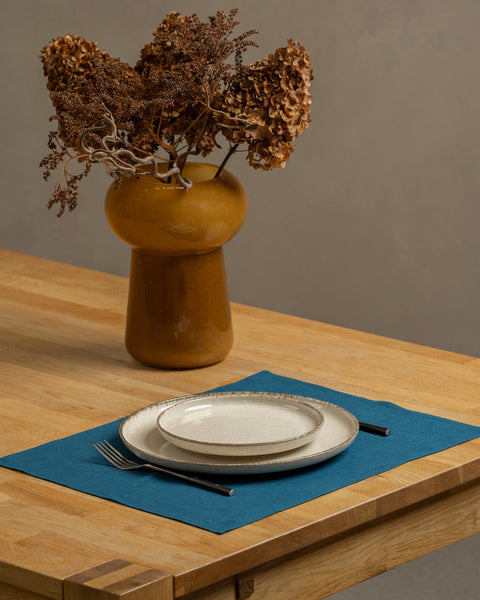 The menique linen placemat in cobalt blue color on a wooden table top. There is a vase on top of the table, as well as some cutlery by the sides of the plate, that is on top of the placemat.
