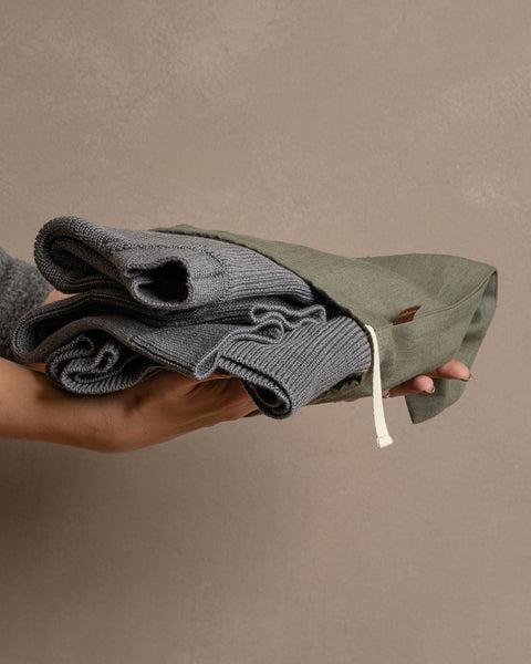 A person's hands gently hold a folded gray knitted garment inside a stone green linen drawstring bag against a muted brown background.