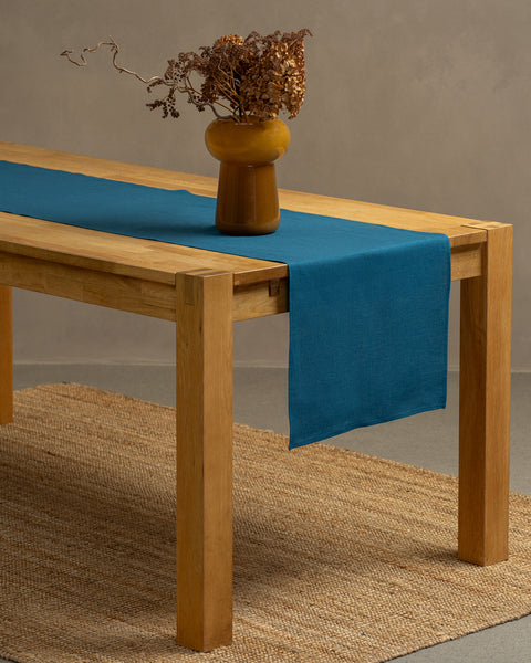 A wooden table positioned on top of a carpet. There is a cobalt blue linen table runner and a vase on top.