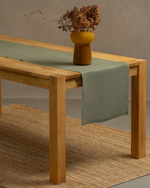 A wooden table positioned on top of a carpet. There is a stone green linen table runner and a vase on top.