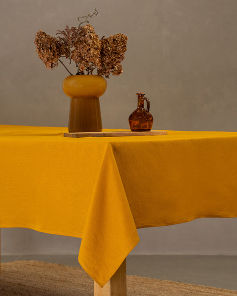 The menique linen tablecloth in spicy yellow color, draping over a wooden table, with a vase, wooden board and a glass bottle on top of it.