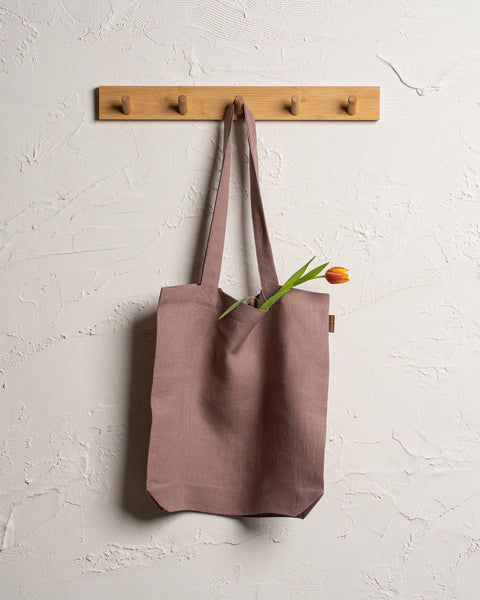 A faded rose reusable linen tote bag hangs from a wooden rack with four pegs mounted on a textured off-white wall. A single tulip with a red and yellow bloom and green leaves is visible sticking out of the top of the bag.