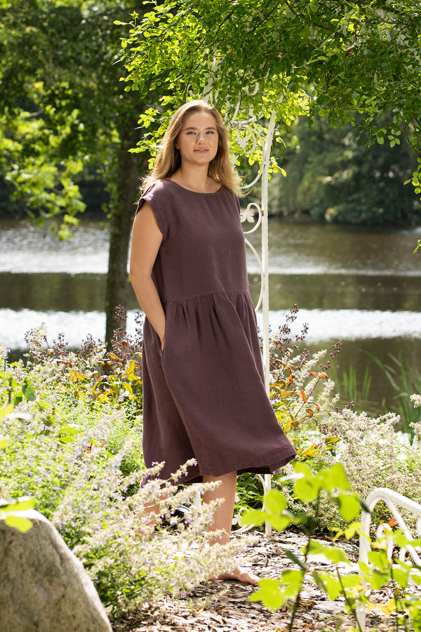 A barefoot woman stands gracefully under a white metal garden archway with a serene lake in the background. She looks directly at the camera, modeling a shadow purple linen dress with a gathered skirt and her hands resting in the side pockets.