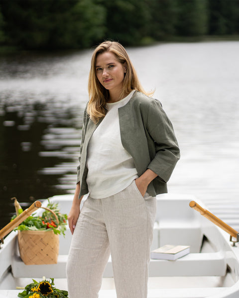 A woman stands confidently inside a small white rowboat on a lake, casually resting one hand in the pocket of her trousers. Her outfit features a stone green linen jacket worn open over a pure white top and loose-fitting natural linen pants.