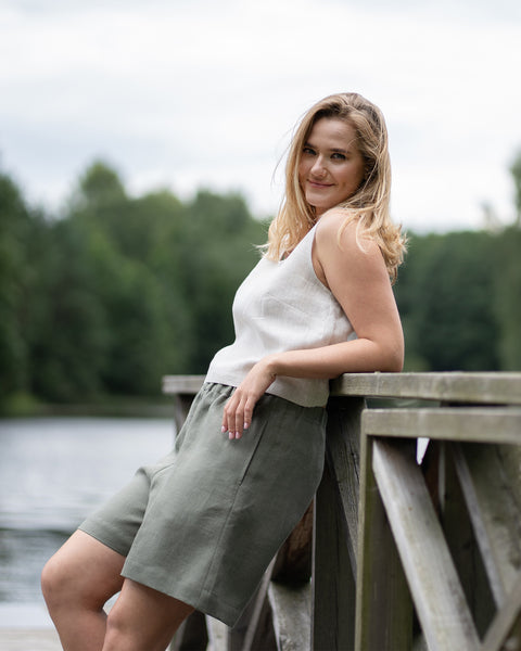 A smiling woman leans comfortably against a wooden pier railing, looking directly at the camera with her hair gently blowing in the wind. She is wearing a natural linen sleeveless top and stone green bermuda shorts, set against a softly blurred background of a lake and green trees.