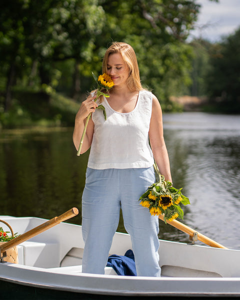 Standing inside a small boat on a lake, a woman closes her eyes to softly smell a bright yellow sunflower. She wears a crisp white linen top and light blue pants while holding a bundle of freshly picked sunflowers at her side.