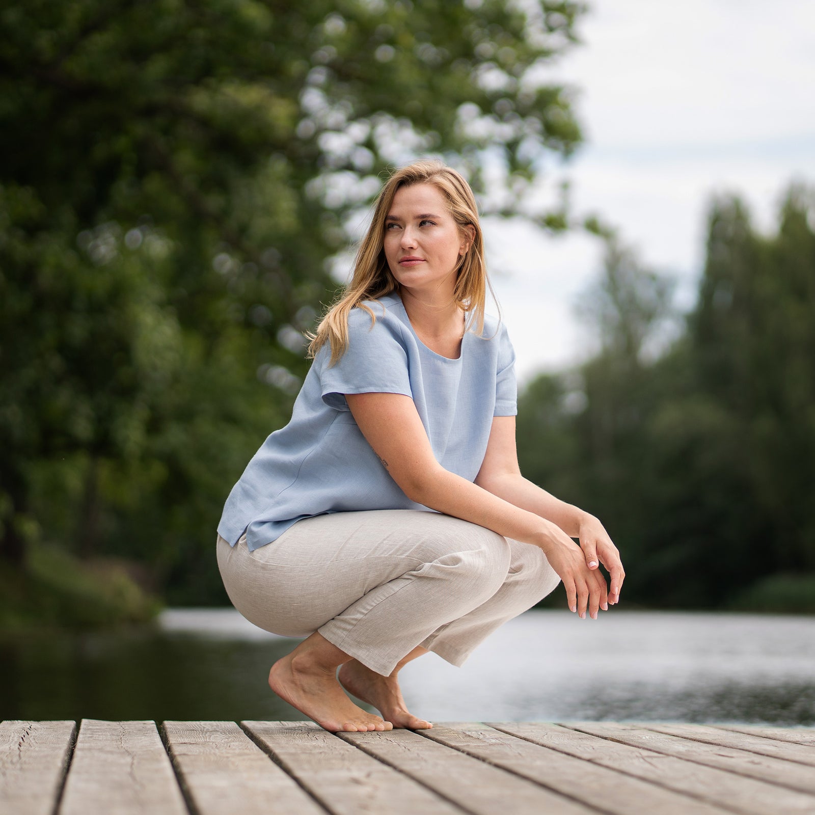 A barefoot woman leans gracefully against a wooden pier railing, gazing off to the side while brushing a hand through her hair. She wears a natural linen top and stone green bermuda shorts, with a peaceful lake stretching out behind her.