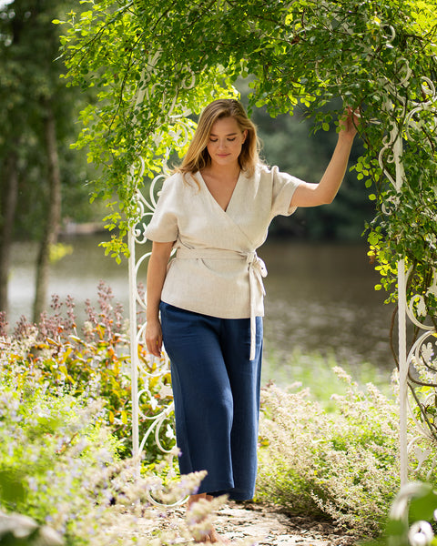 A barefoot woman pauses under a leafy garden archway near a lake, looking downward with a serene expression. Her outfit features a natural-toned linen wrap top secured with a waist tie, complemented by wide-leg, storm blue linen pants.