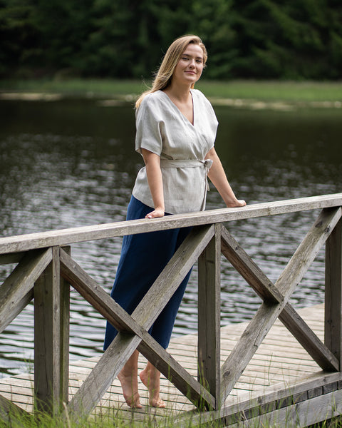 A woman stands barefoot on a wooden lakeside pier, resting her hands lightly on the diagonal railing and smiling at the camera. She is wearing a natural-colored, short-sleeve linen wrap top and loose-fitting, storm blue linen pants.