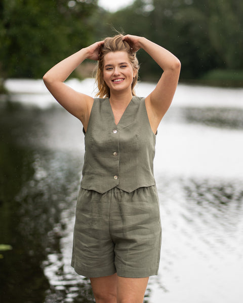 A woman stands by a tranquil lake, smiling brightly at the camera as she runs both hands through her blonde hair. She is wearing a coordinated stone green linen outfit consisting of a button-up vest and matching shorts.